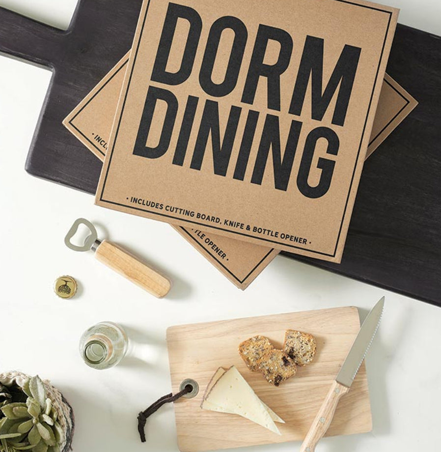 A dorm room dining kit including a brown cutting board, a knife, a bottle opener, and a cheese board, packaged in a box with 'DORM DINING' printed on it.